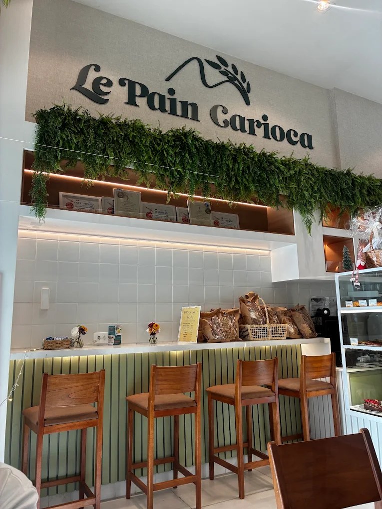 Salão da Le Pain Carioca: balcão verde com banquetas de madeira, samambaias e a logo da casa na parede